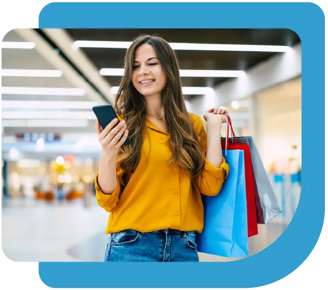 Smiling young woman in a yellow blouse holding shopping bags and looking at her smartphone in a mall.