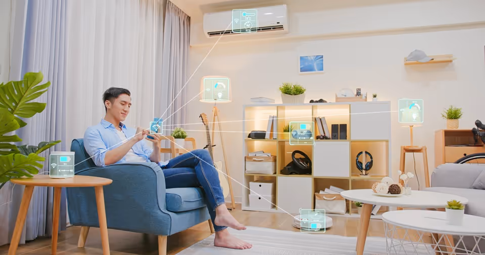 Man sitting on a blue armchair using a smartphone in a modern living room with smart home device icons overlaid indicating connected lighting, air conditioning, security, and robot vacuum.