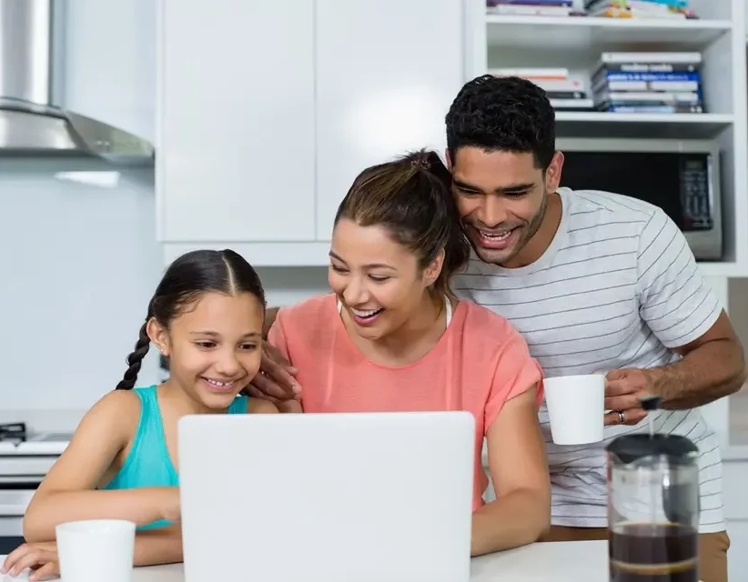 Smiling family of three gathered around a laptop in a kitchen, with the father holding a white mug.