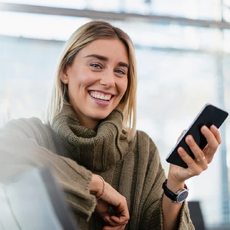 Smiling woman wearing a brown sweater holding a smartphone indoors.