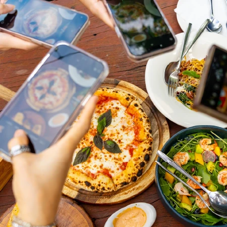 People taking photos of a margherita pizza, pasta, and shrimp salad on a wooden table.