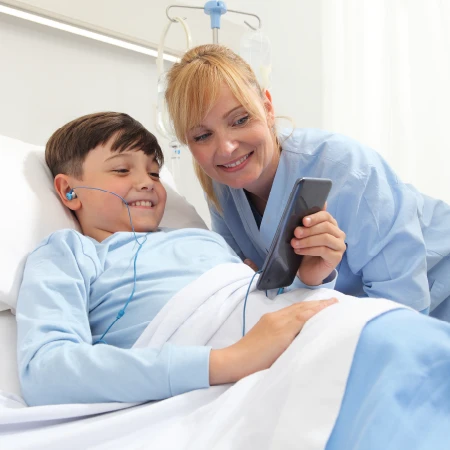 Smiling nurse and boy patient in hospital bed looking at a smartphone together, the boy wearing earphones.
