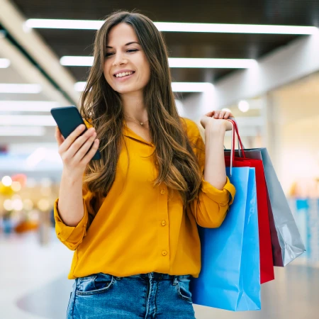 Smiling woman in a yellow blouse holding shopping bags and looking at her phone in a mall.