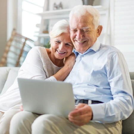 Smiling elderly couple sitting on a couch looking at a laptop together in a bright living room.