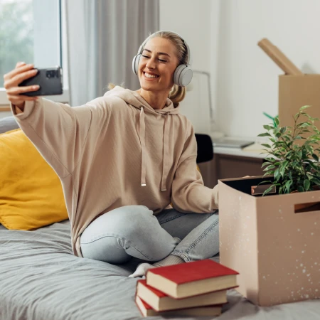 Smiling young woman with headphones taking a selfie while sitting on a bed surrounded by books and a moving box with a plant.
