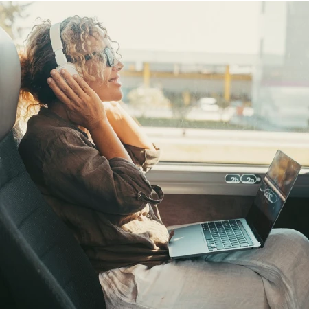 Woman with curly hair wearing headphones, sitting on a train seat with a laptop on her lap, looking out the window.