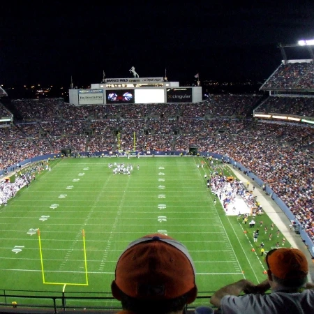 Nighttime view of a packed football stadium with players huddled on the field and spectators in the stands.