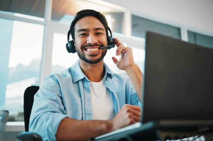 Smiling man wearing headset and sitting at desk with laptop in bright office.