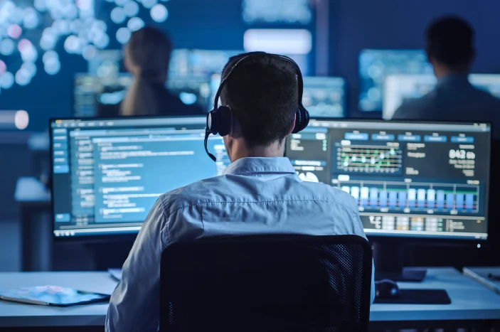 Person wearing headset working on dual monitors displaying data and code in a dark control room with two other people in background.