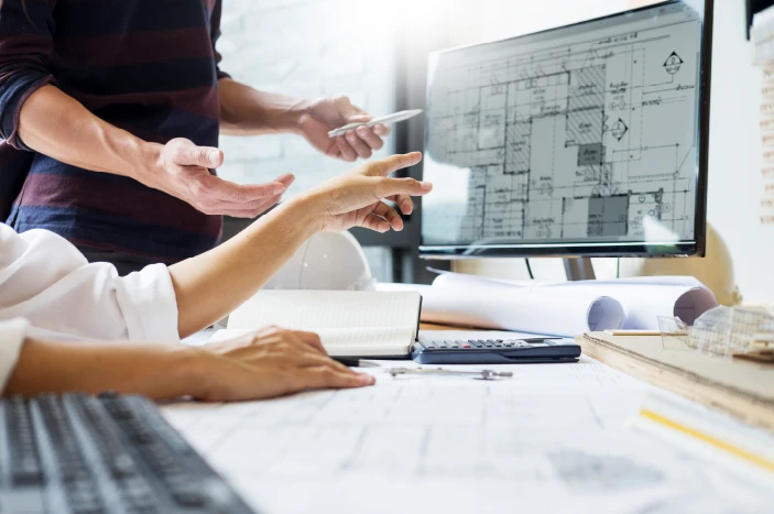 Two people discussing architectural blueprints displayed on a computer monitor, with additional documents and a calculator on the desk.