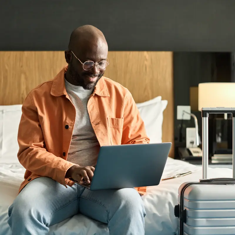 Man wearing glasses and an orange jacket sitting on a hotel bed using a laptop with a silver suitcase nearby.