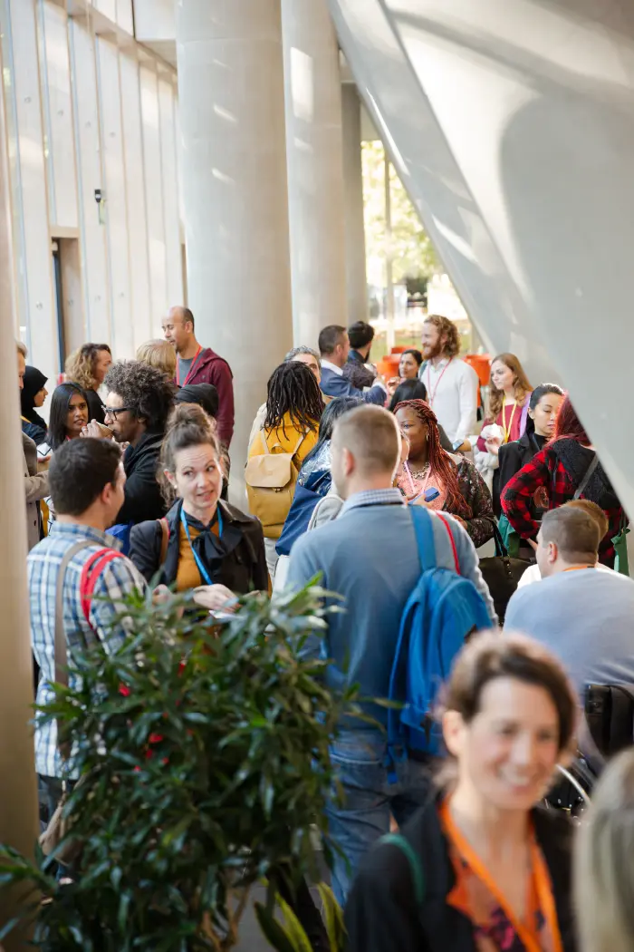 Diverse group of people talking and mingling inside a modern building with large windows and concrete columns.