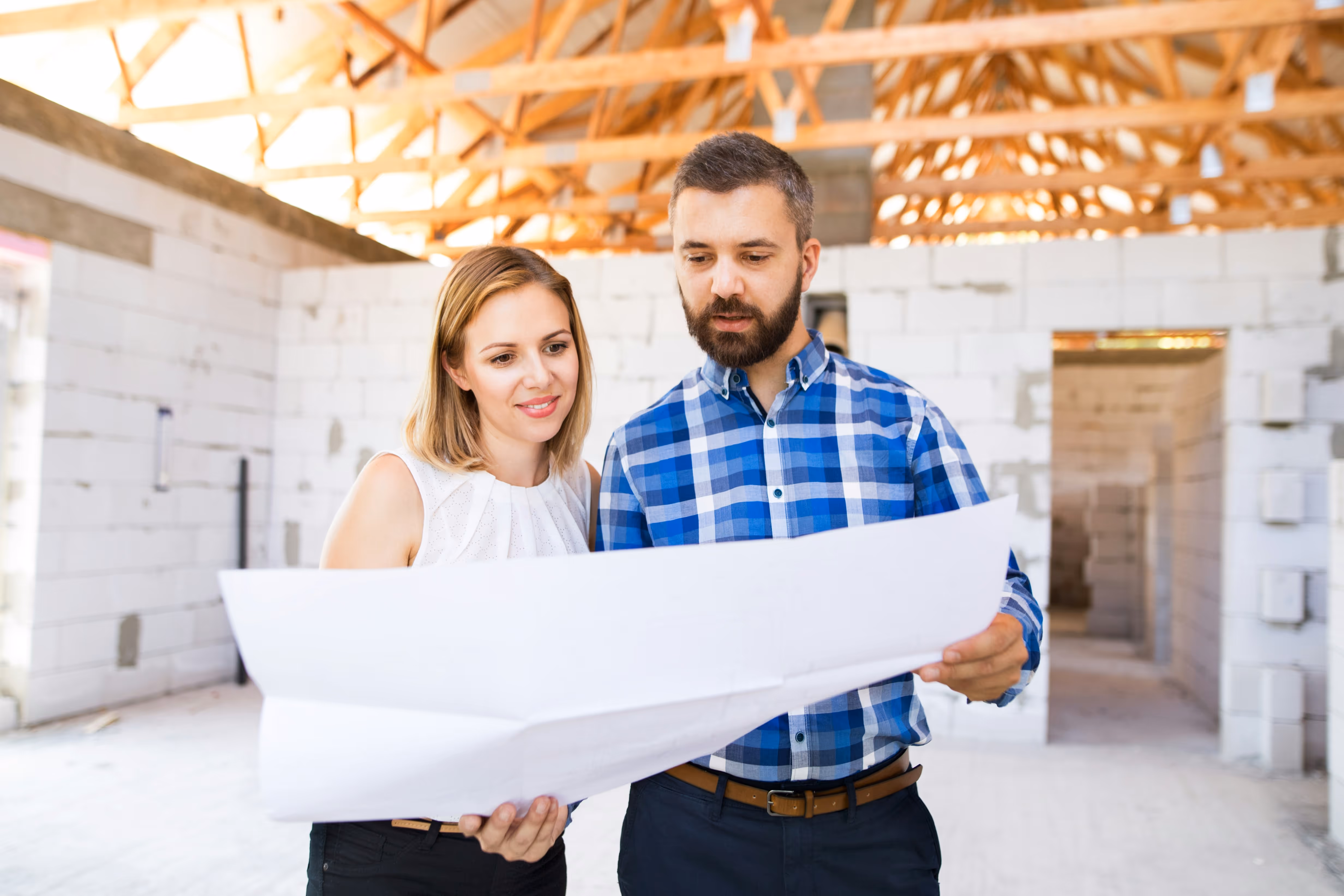 Man and woman reviewing architectural plans inside a building under construction with exposed wooden ceiling beams.