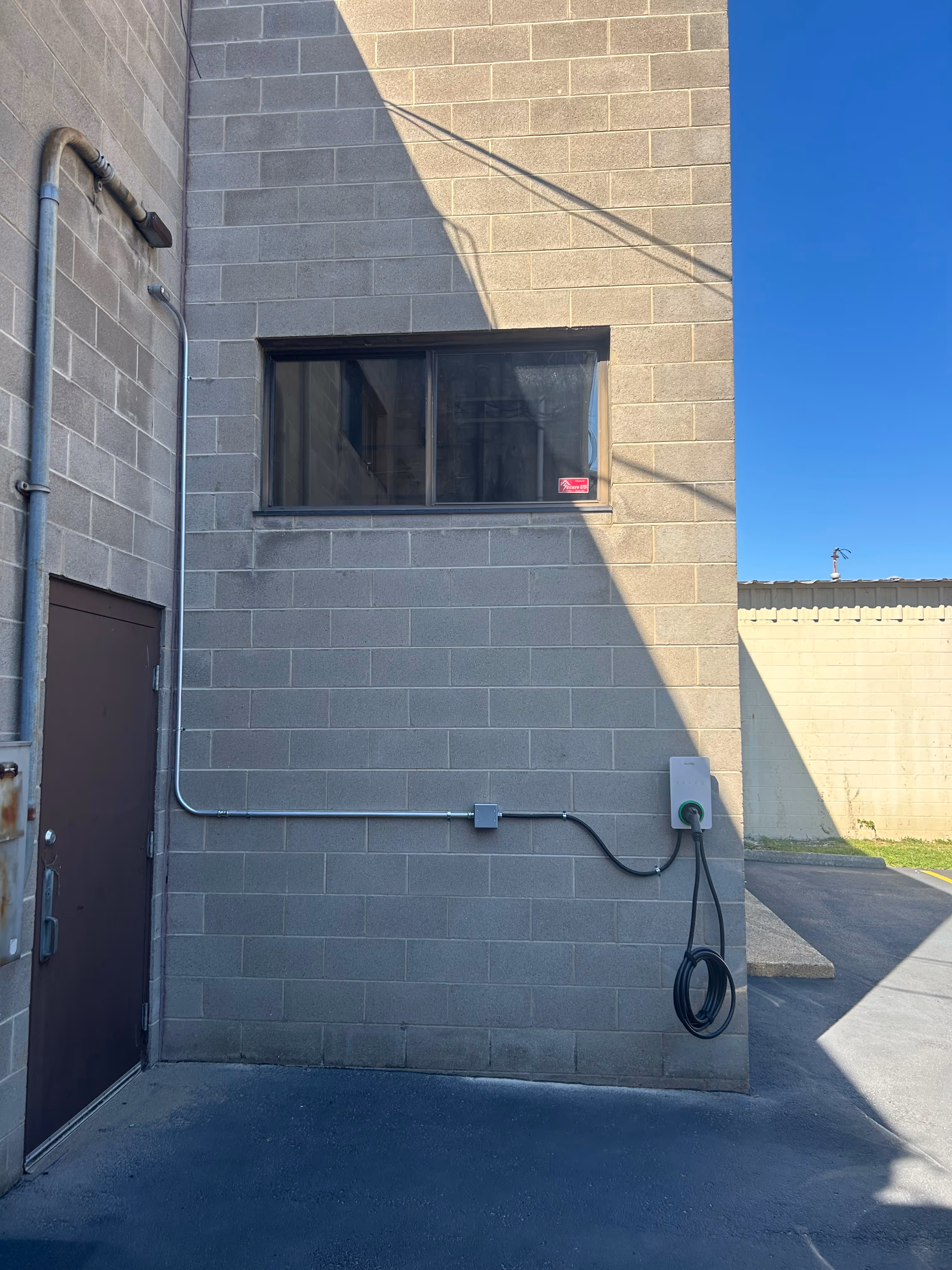 Electric vehicle charging station mounted on a gray cinder block wall next to a closed brown door under a window on a sunny day.