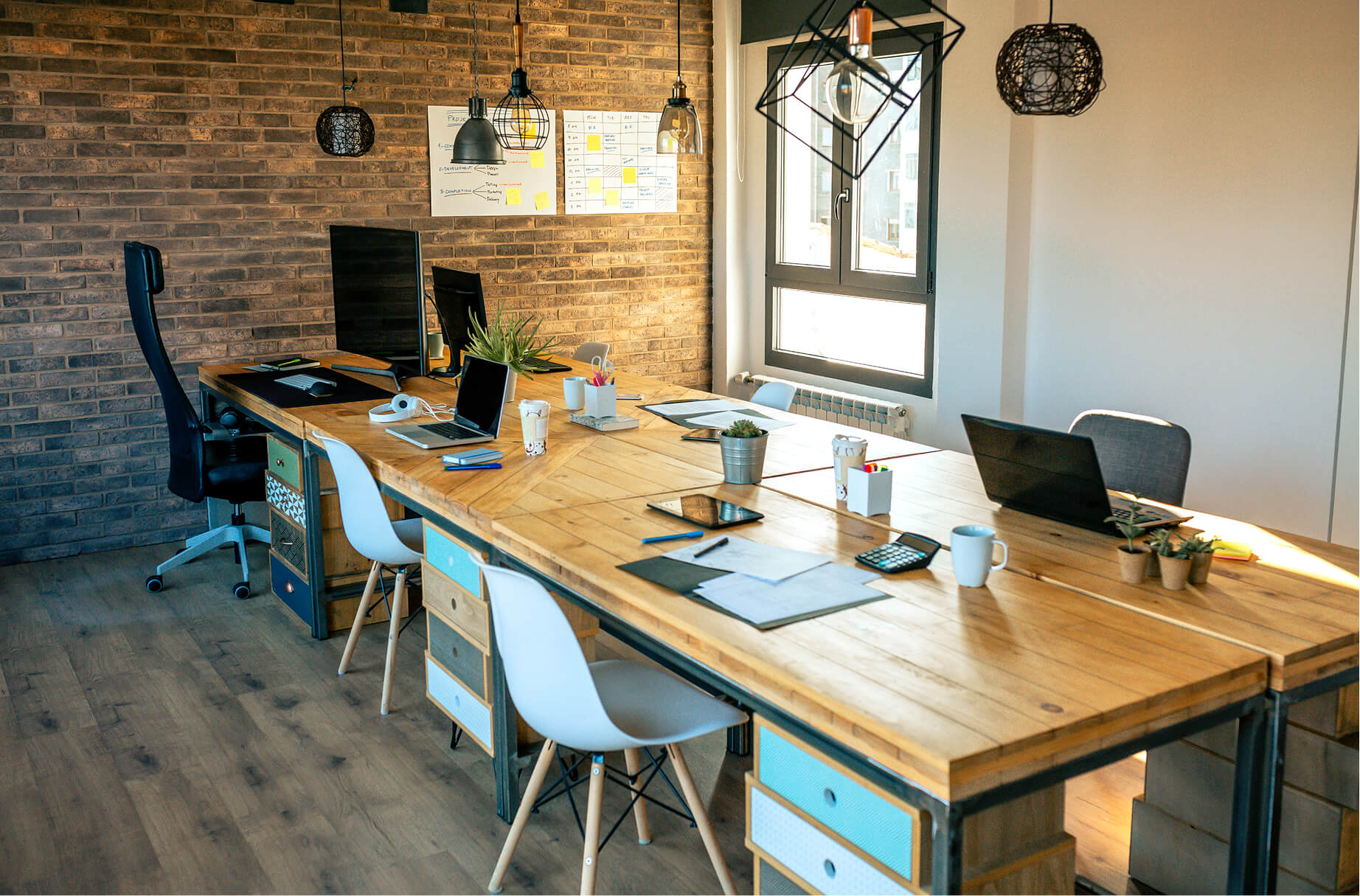 Modern office workspace with wooden desk, computers, plants, and white chairs near brick wall and window.
