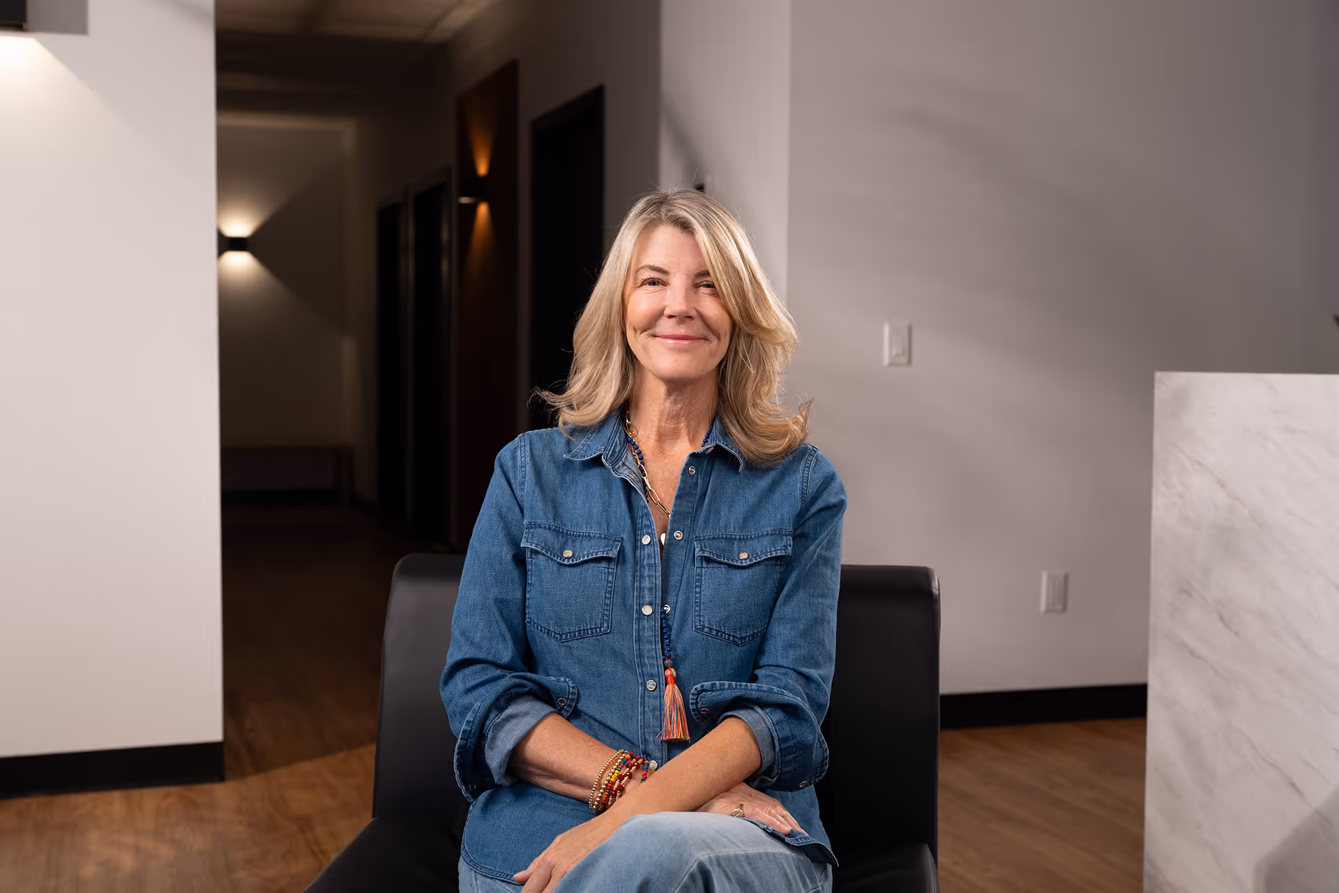Smiling middle-aged woman with blonde hair wearing a denim shirt and colorful bracelets seated indoors on a black chair.