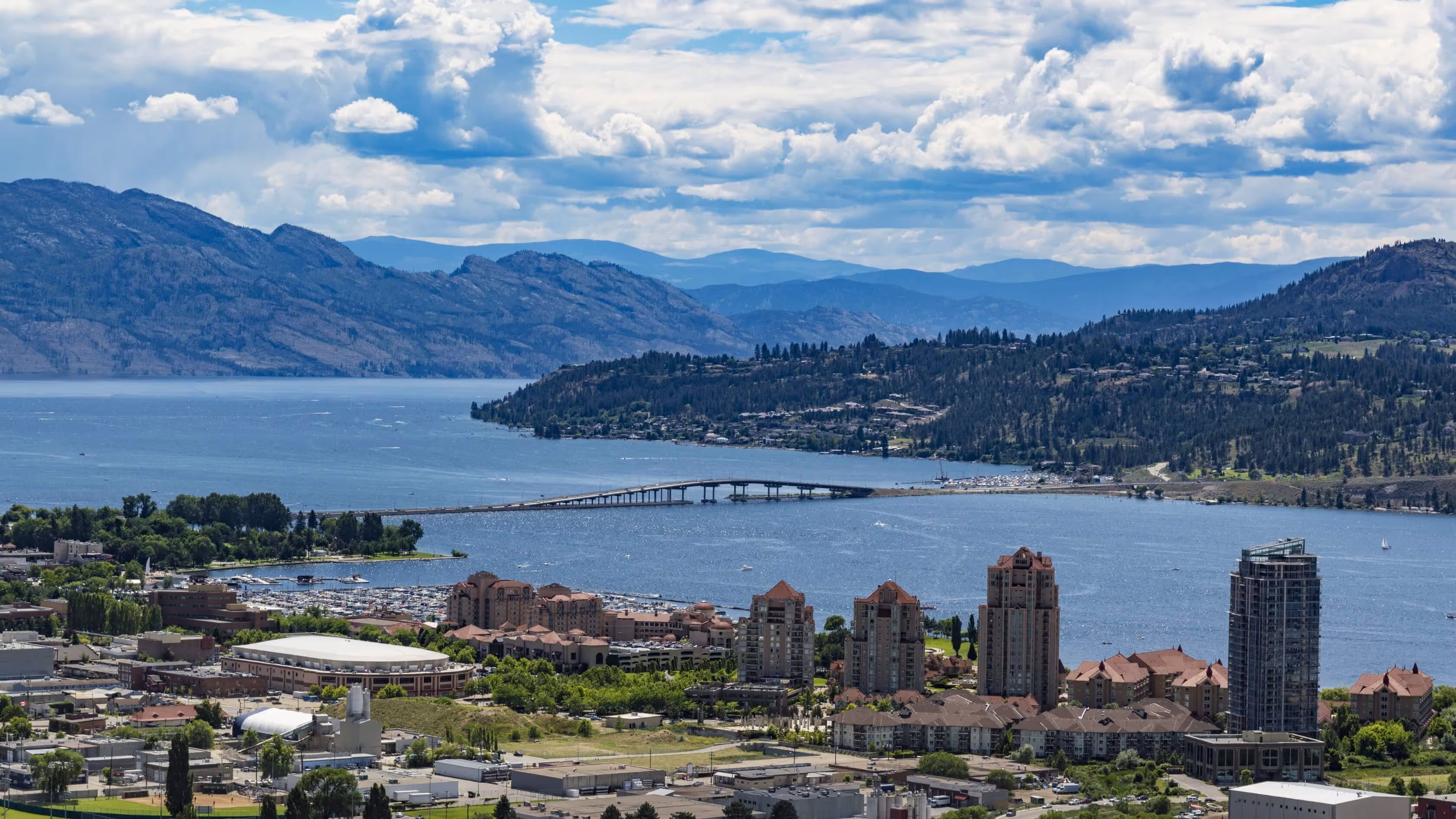 Cityscape with tall buildings near a large lake bordered by mountainous terrain under a partly cloudy sky.