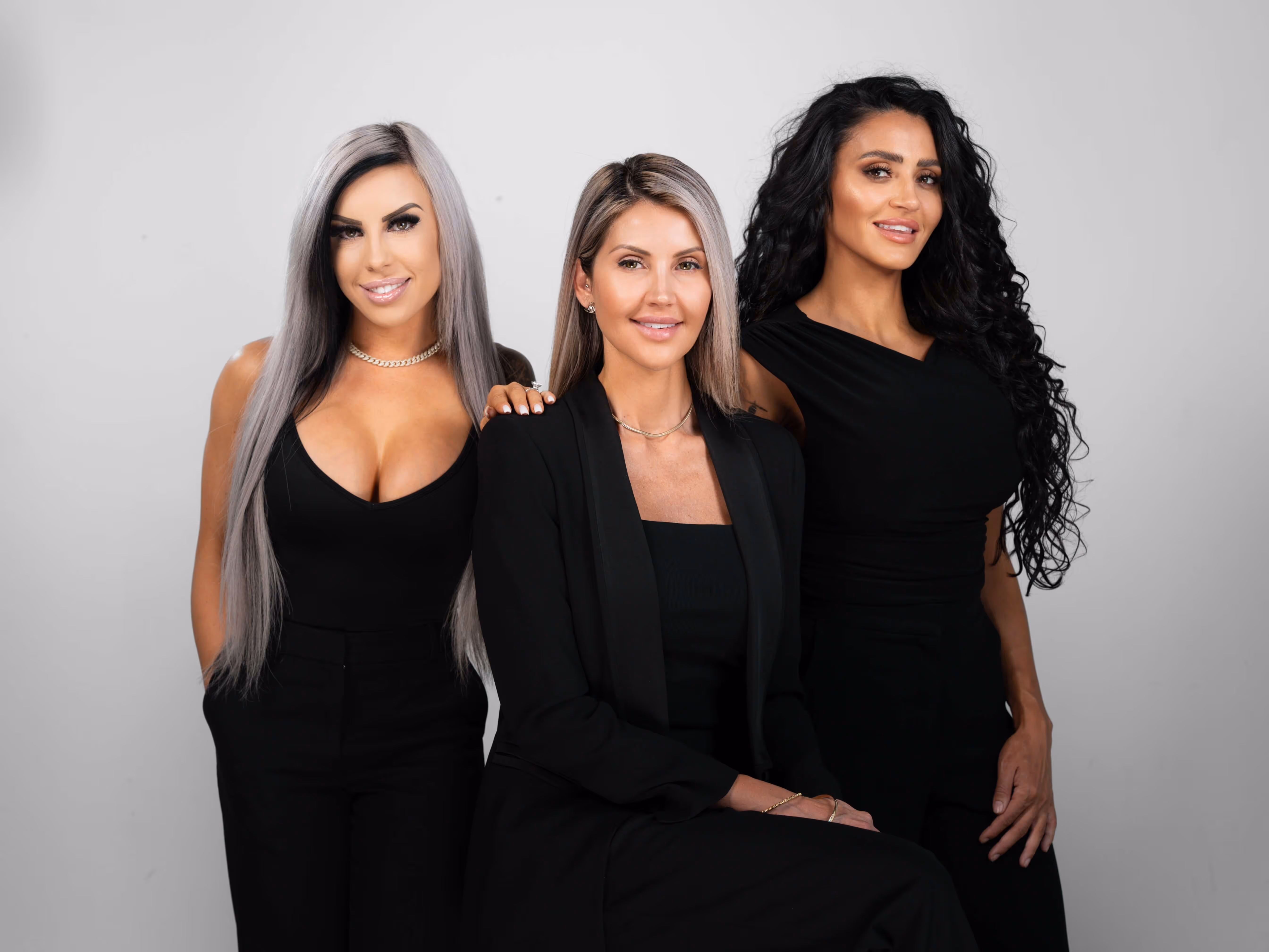 Three women in black outfits posing against a plain light background, two standing and one seated in the center.