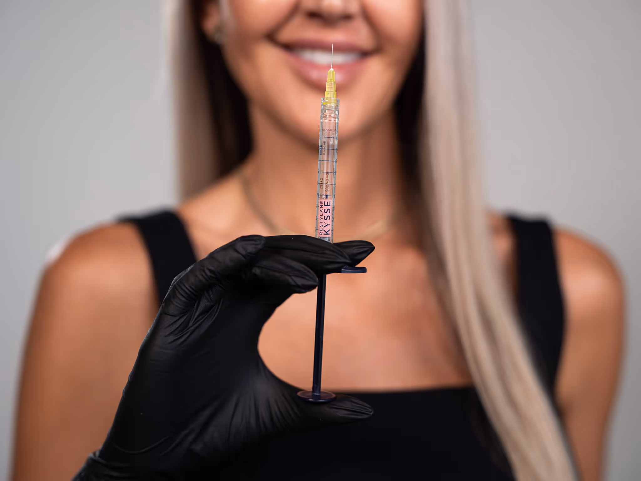 Woman with long blonde hair wearing black gloves, holding a syringe labeled Restylane Kysse.