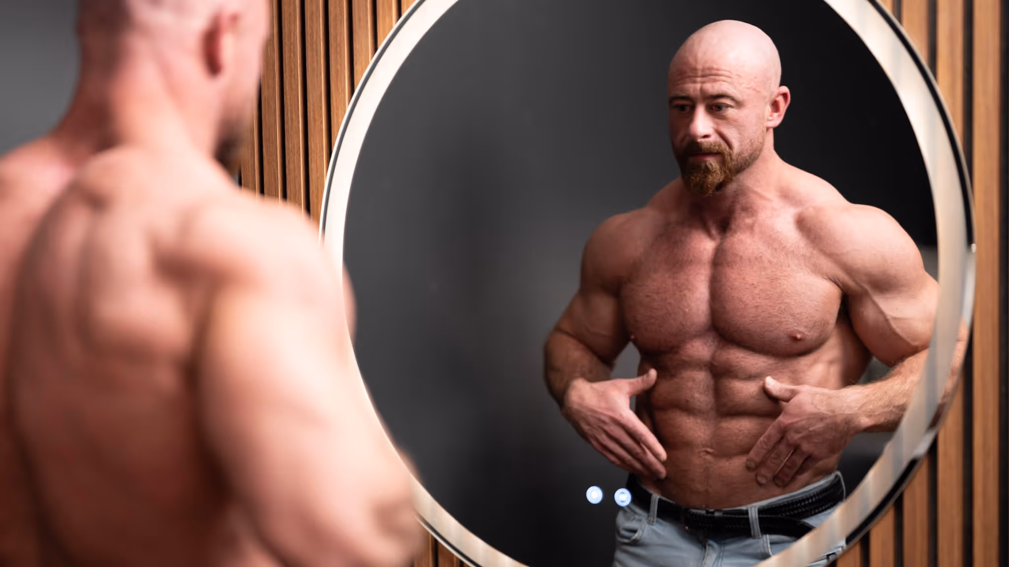 Muscular bald man with a beard looking at his toned torso in a round mirror against a wooden wall.