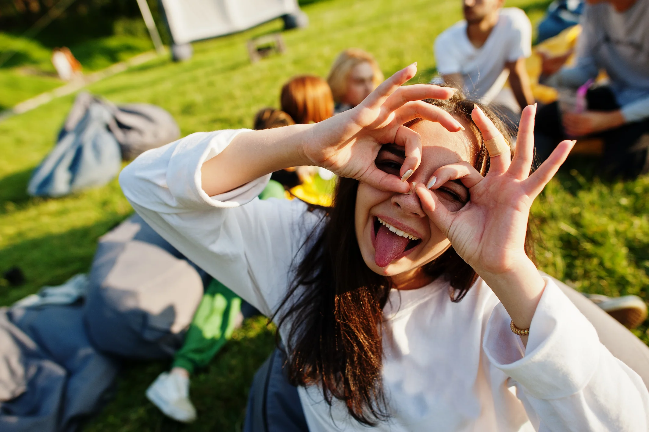 Young woman outdoors making glasses shape with hands over her eyes and sticking out her tongue, with friends sitting on the grass in the background.