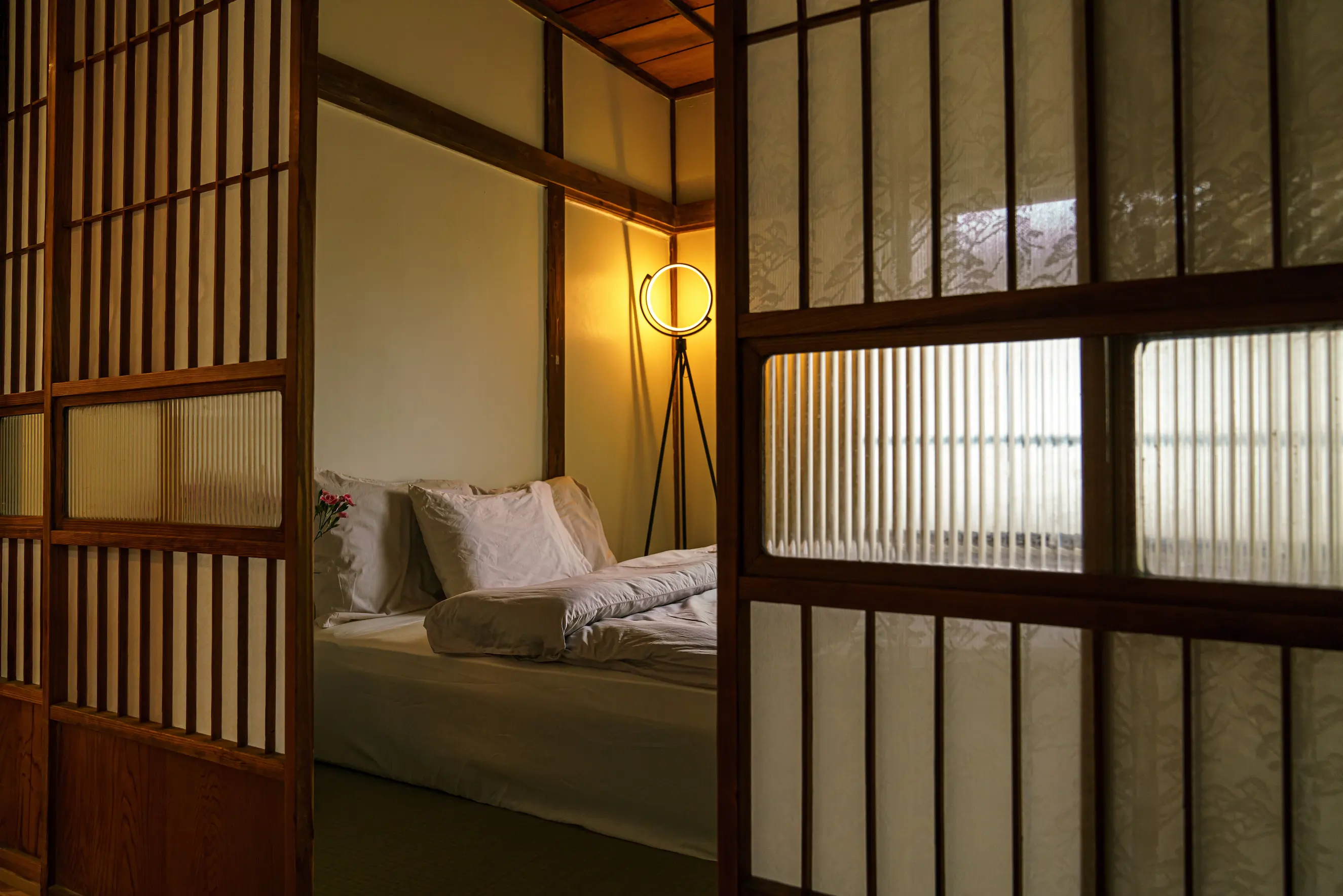 Cozy bedroom with white bed linens, a floor lamp with circular light, and wooden sliding doors partially open.