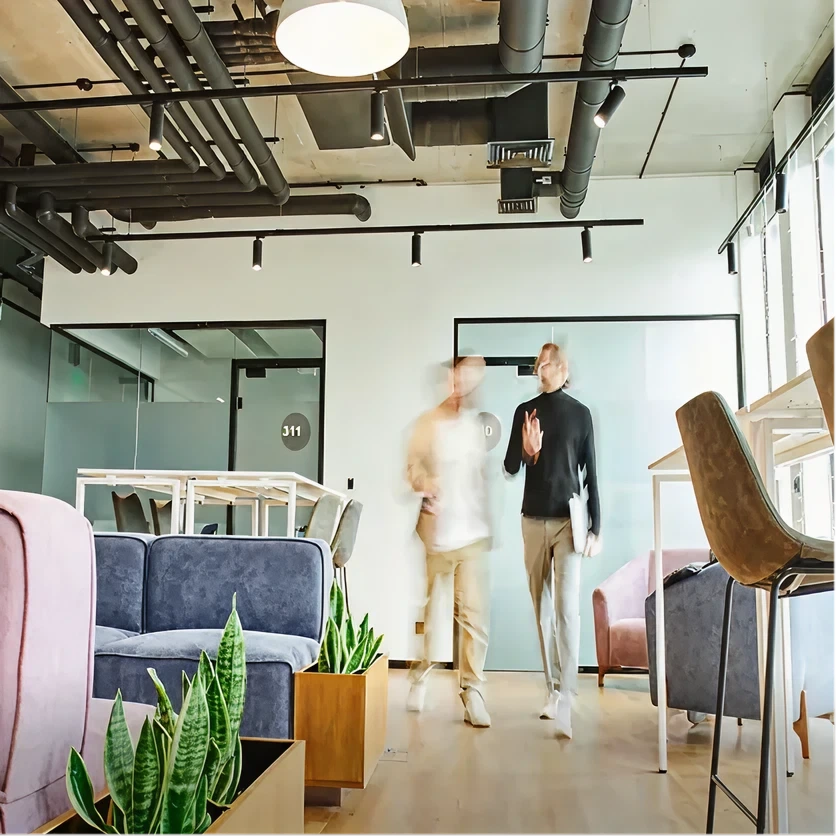 Two blurred figures walking in a modern office lounge area with sofas, plants, and large windows.