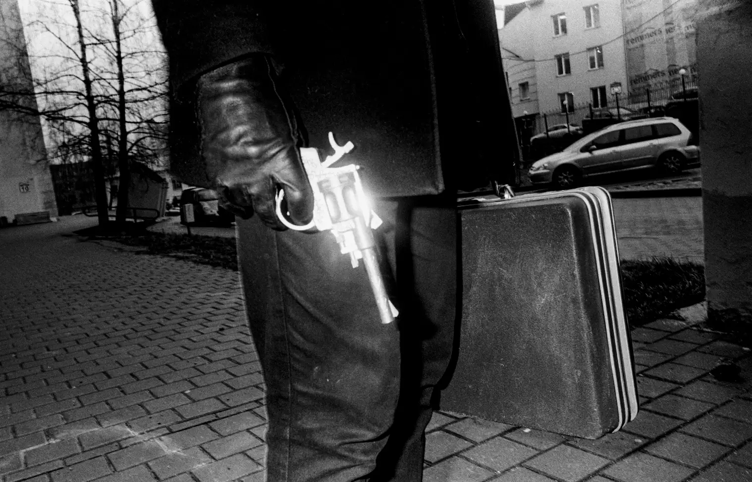 Person wearing black gloves holding a revolver and a dark briefcase, standing on a paved street with buildings and parked cars in the background.