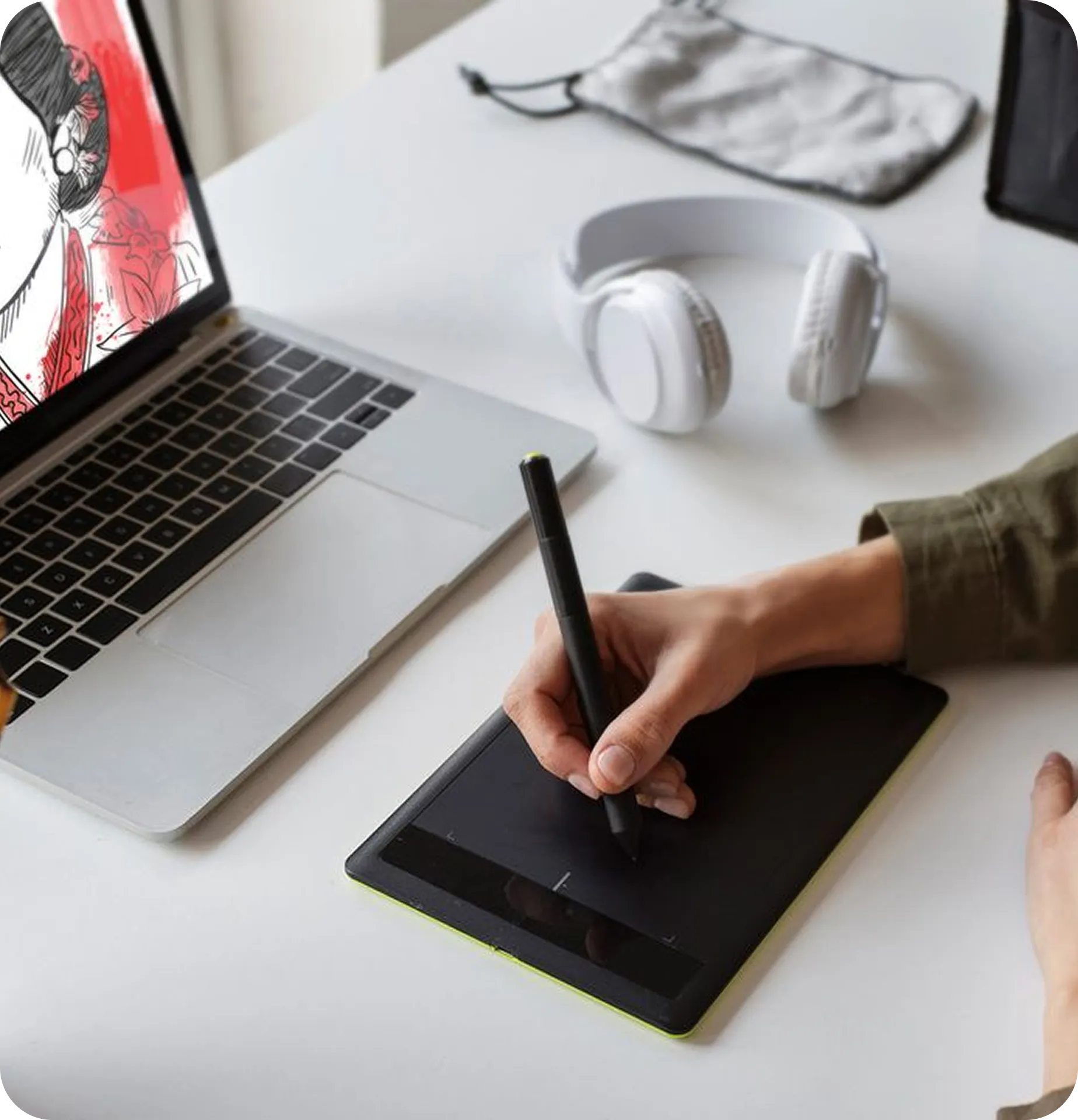 Hand, das einen Stift auf einem Grafiktablet hält neben einem Laptop und weißen Kopfhörern auf einem weißen Schreibtisch.