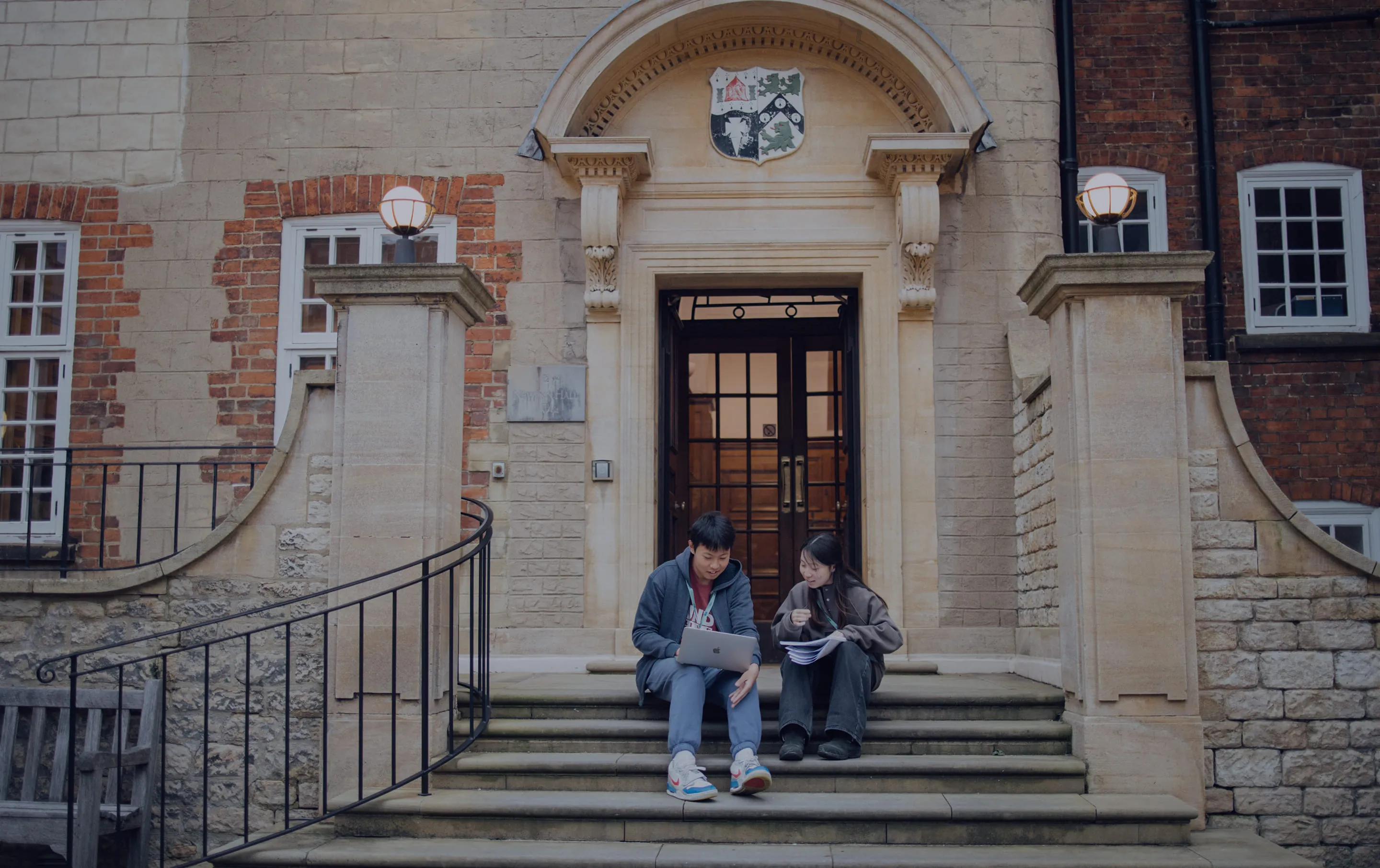 Two students sitting on steps outside a historic building, studying with a laptop and papers.