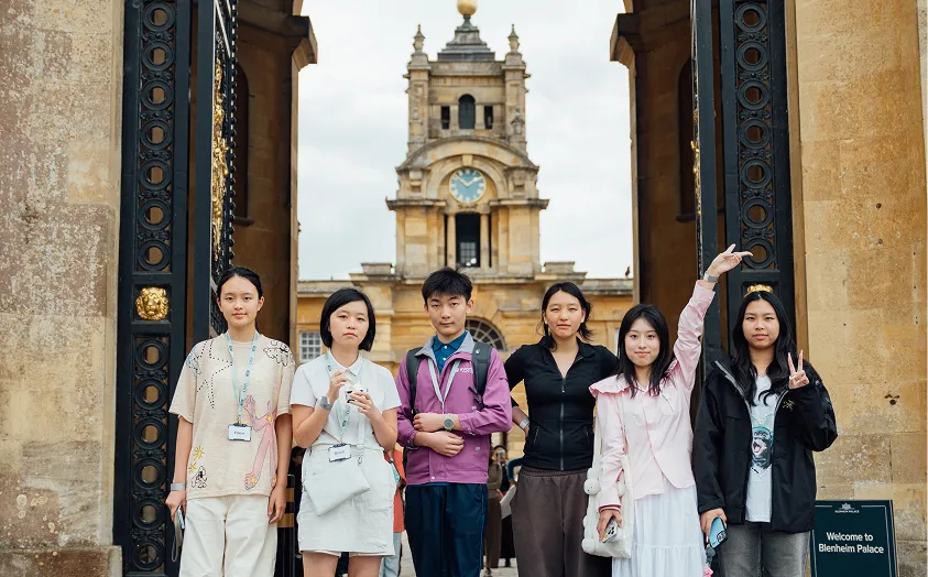 Group of six young people posing in front of ornate gates and clock tower at Blenheim Palace.