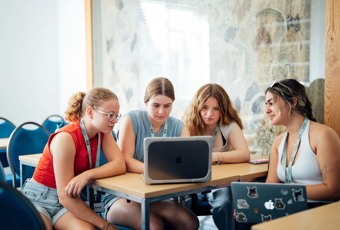 Four young women intently looking at a laptop on a table in a classroom with blue chairs.
