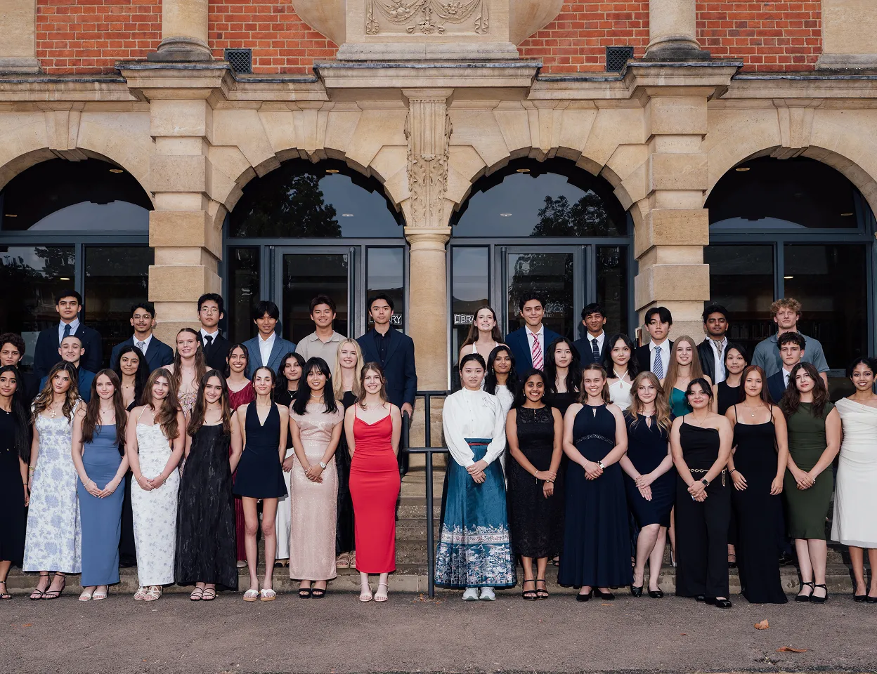 Large diverse group of young adults in formal attire posing outside a brick building with arched windows.