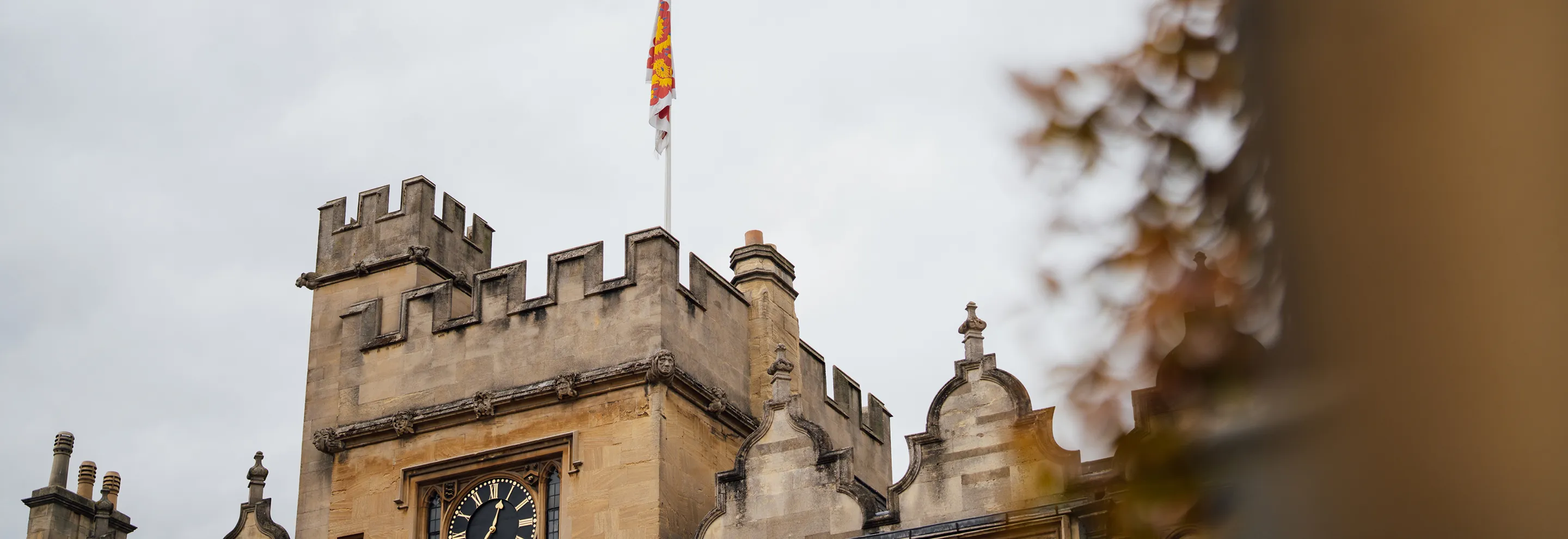 Stone clock tower with battlements, chimneys, and a flag on a cloudy day.