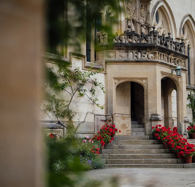 Stone building entrance with stairs lined by red flowers in pots and stone carvings above doorway.