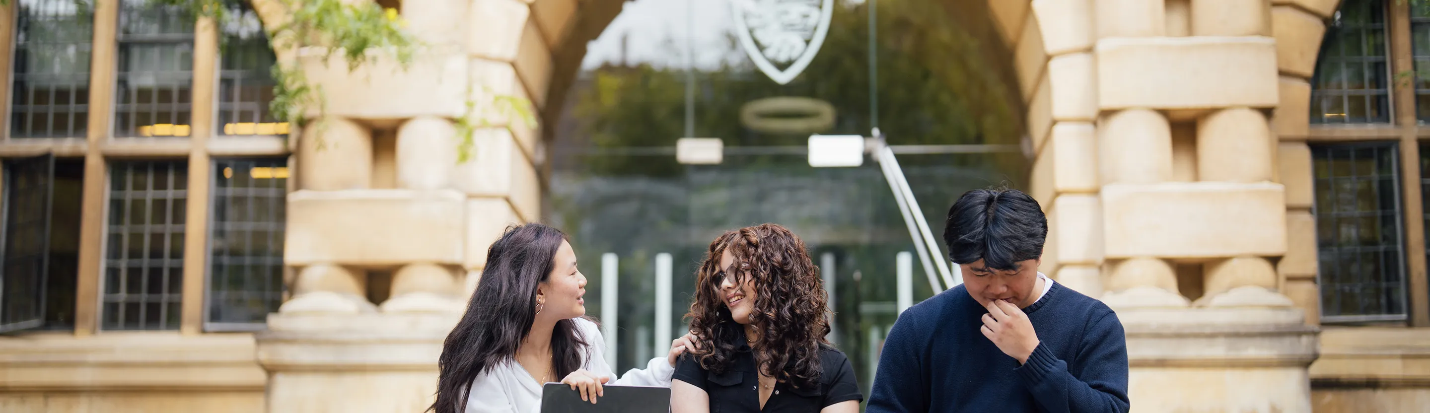 Three students sitting outdoors in front of a university building, interacting and smiling.