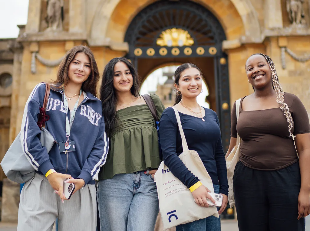 Four diverse young women standing smiling outdoors in front of a historic stone archway.