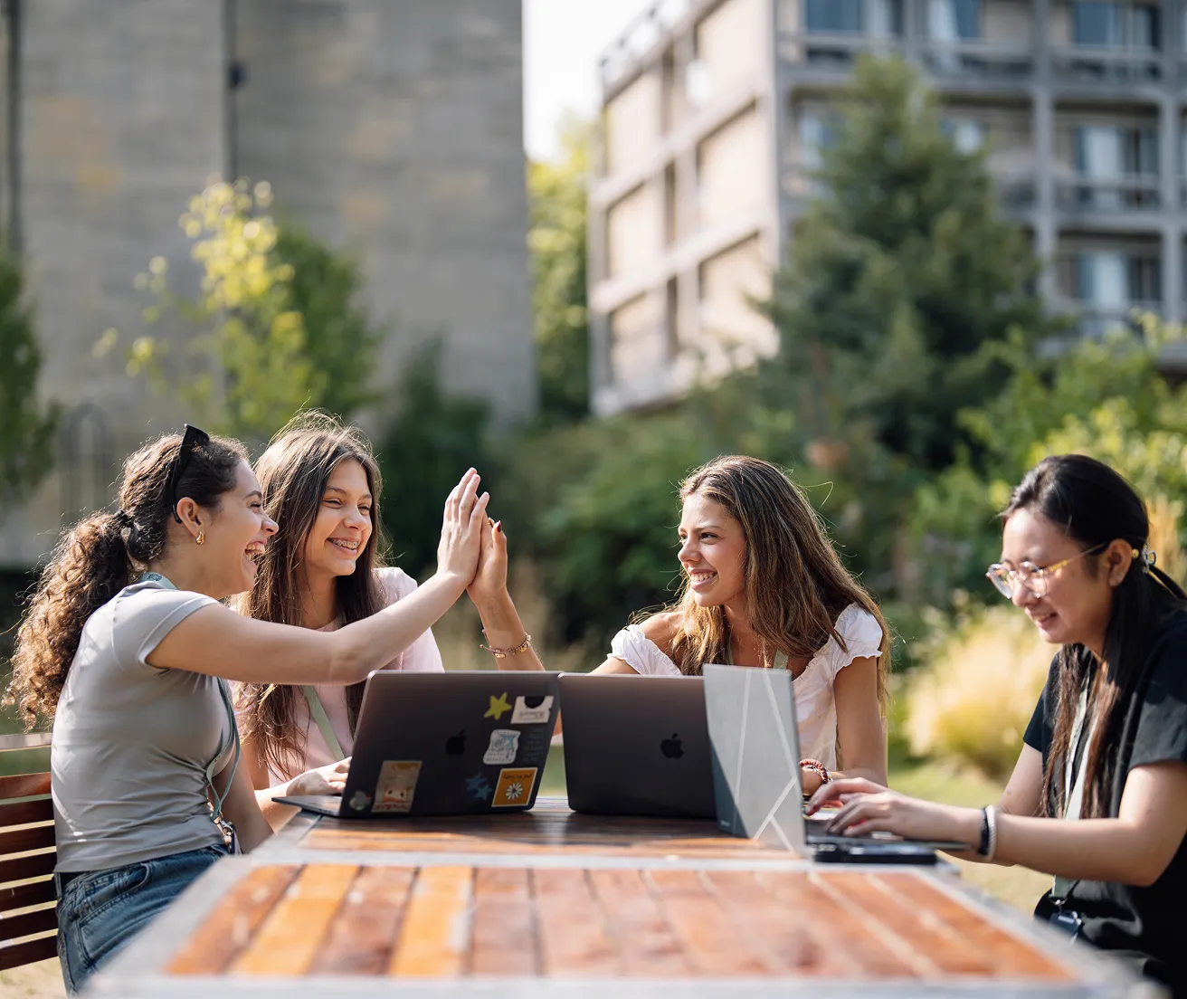 Four young women working on laptops outdoors; two friends high-five while the others work nearby.