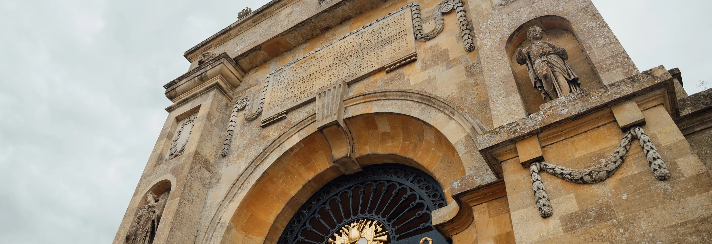Stone archway with decorative statues and engraved plaque under cloudy sky.