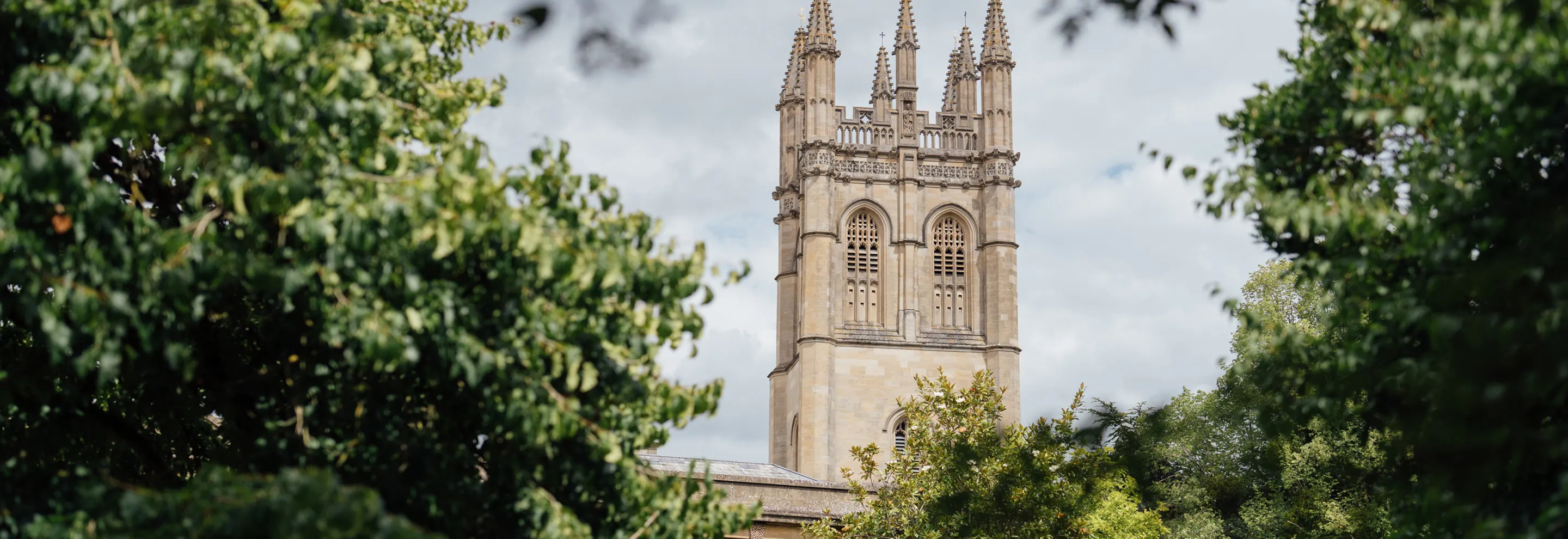 Gothic-style stone tower with spires framed by green trees under a cloudy sky.