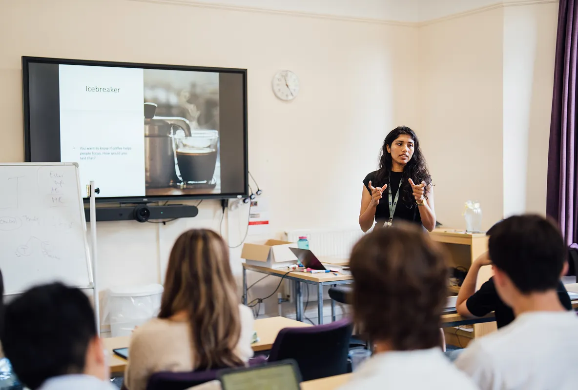 Woman presenting an icebreaker slide about coffee steaming to seated audience in classroom.