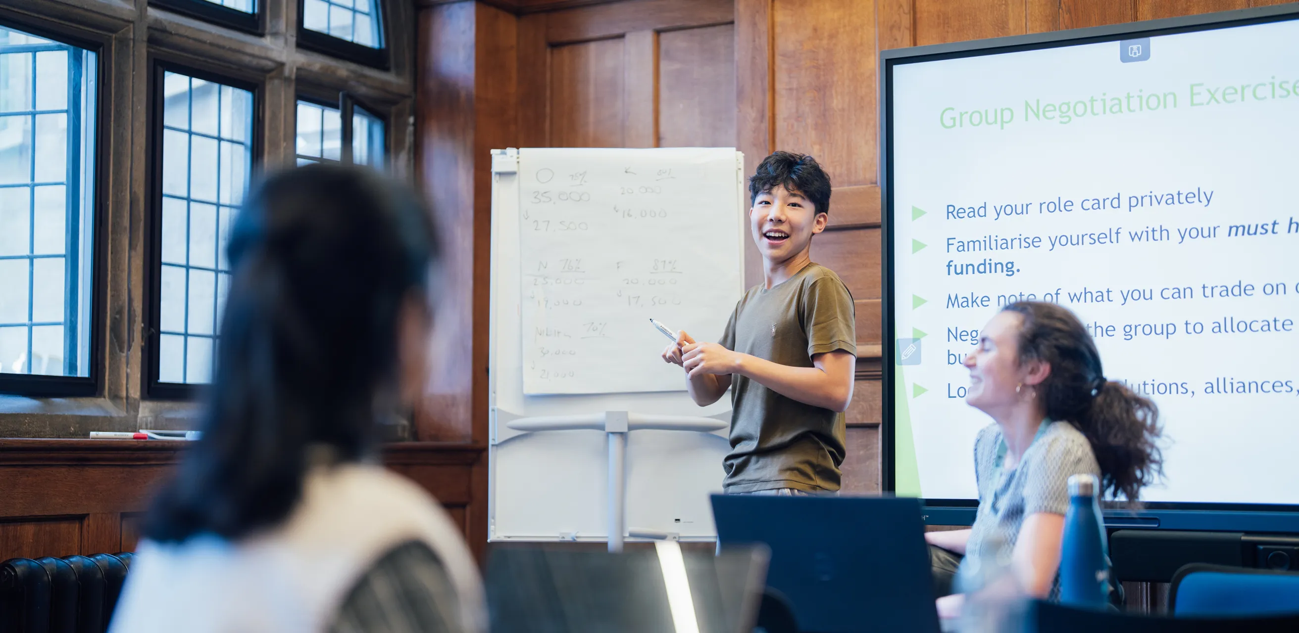 Young man presenting in a room with wooden walls, a flip chart, and a group negotiation exercise on screen.