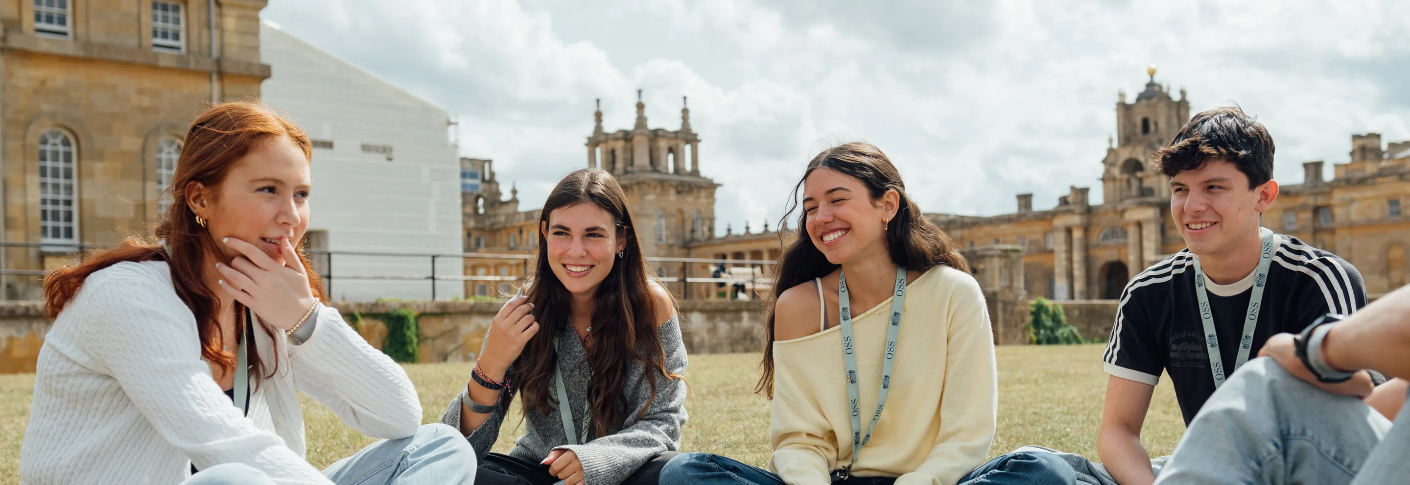 Four young adults sitting on grass outside, smiling and talking in front of historic buildings.