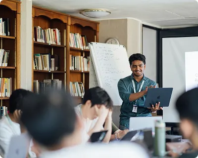 Man holding a laptop, presenting in a classroom with a whiteboard and seated students.