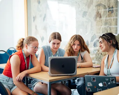 Four young women intently looking at two laptops on a table in a well-lit room.