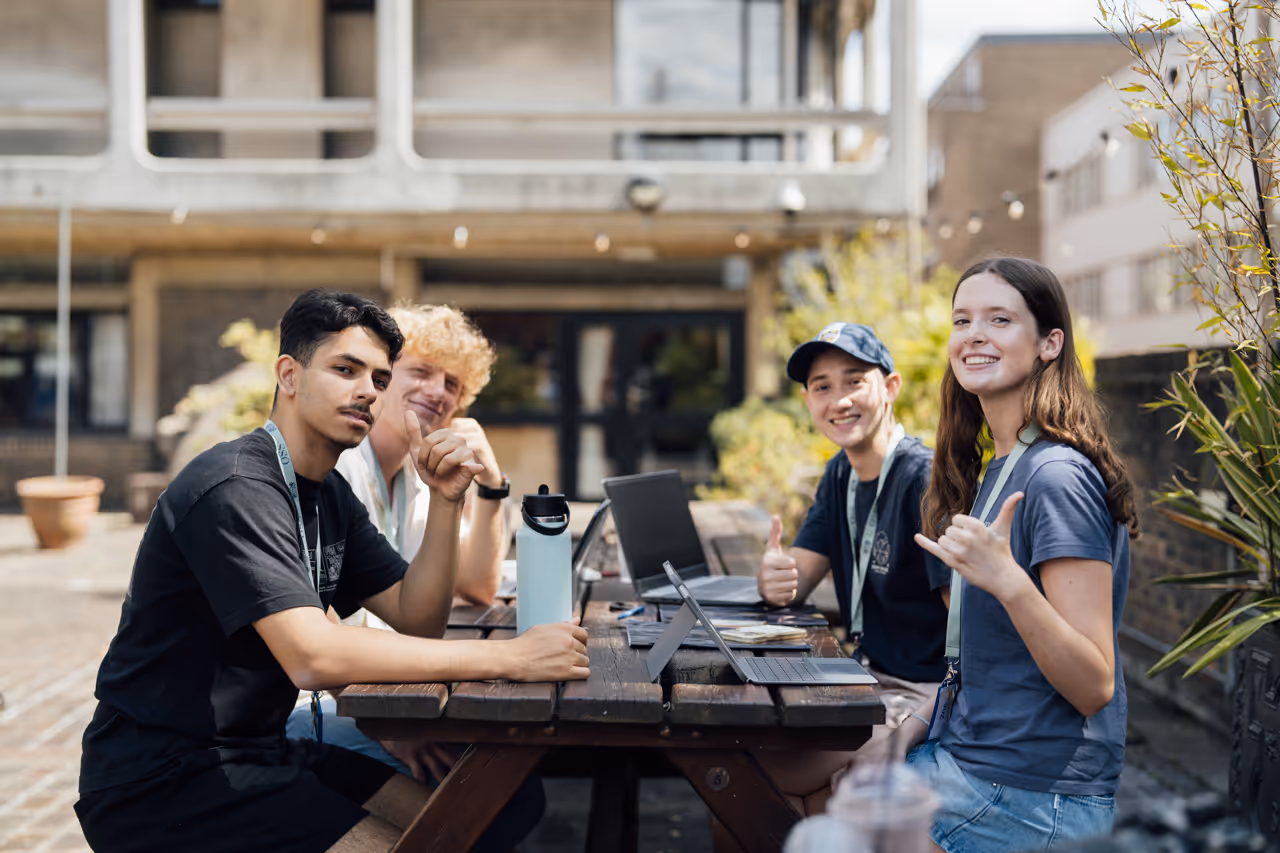 Four young adults sitting outdoors at a wooden table with laptops, smiling and giving thumbs-up gestures.