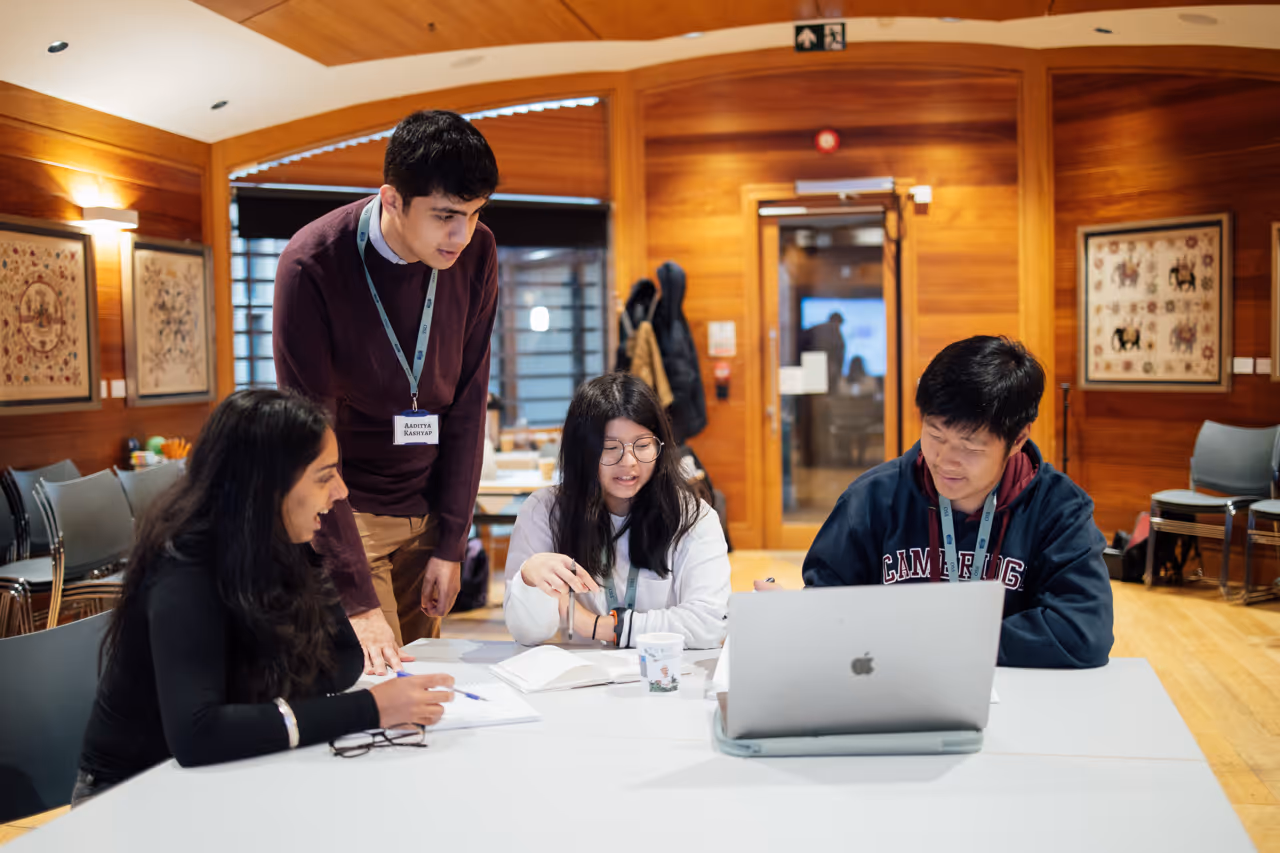 Four students collaborating around a table with laptop and notebooks in a wood-paneled room.