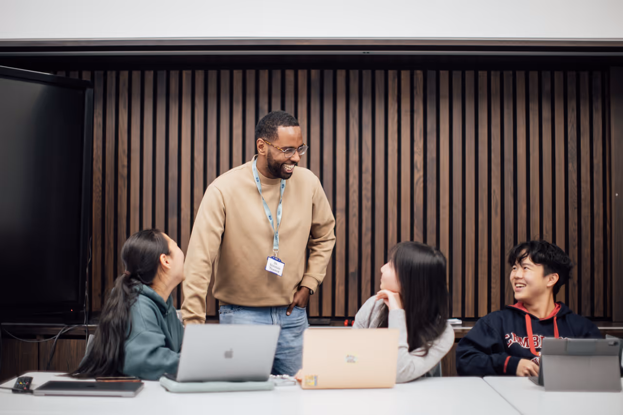 Man standing and smiling while talking to three seated students with laptops and tablets.