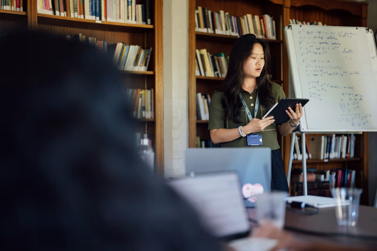 Woman in library presenting with tablet near whiteboard covered in mathematical formulas.