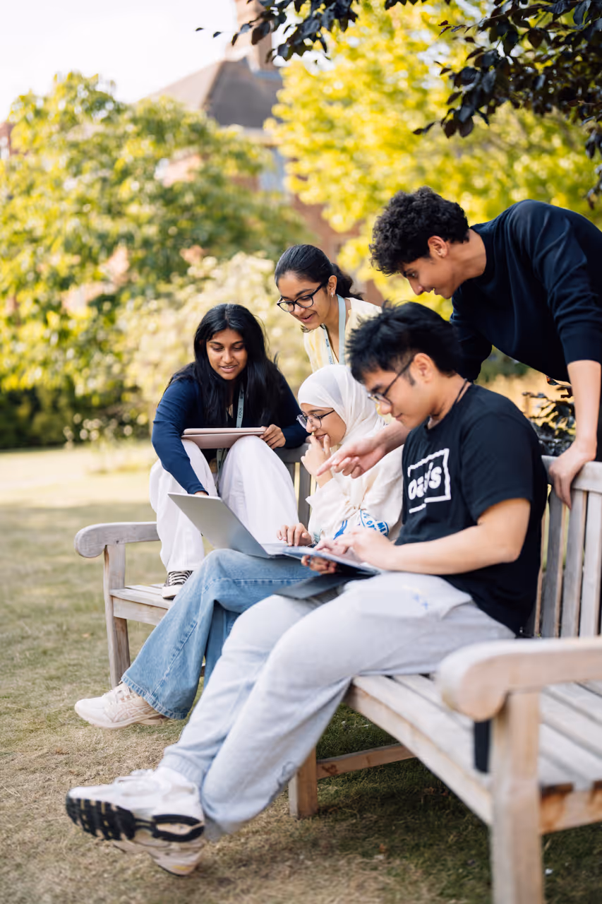 Four diverse young adults studying together outside on a bench with laptops and notes.